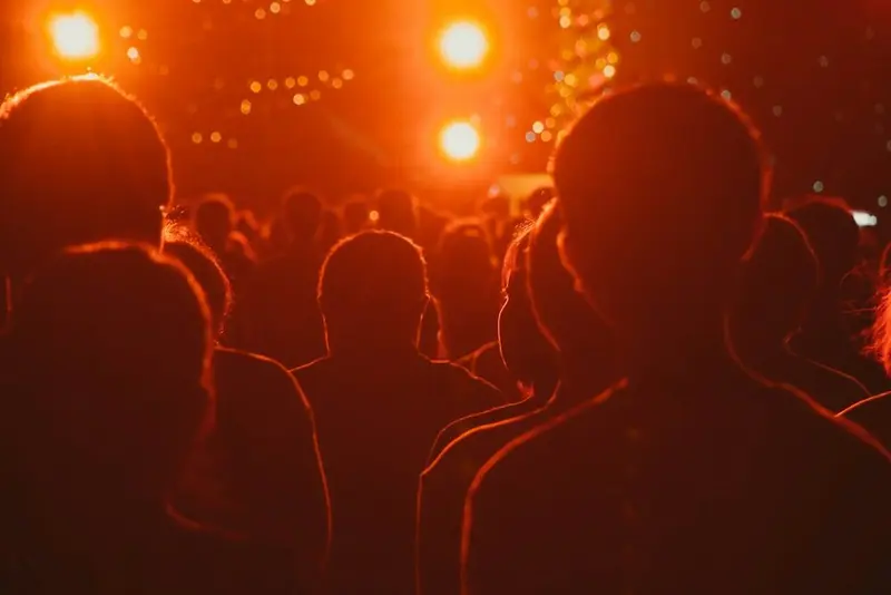 An audience in silhouette from behind, warm orange stage light and haze