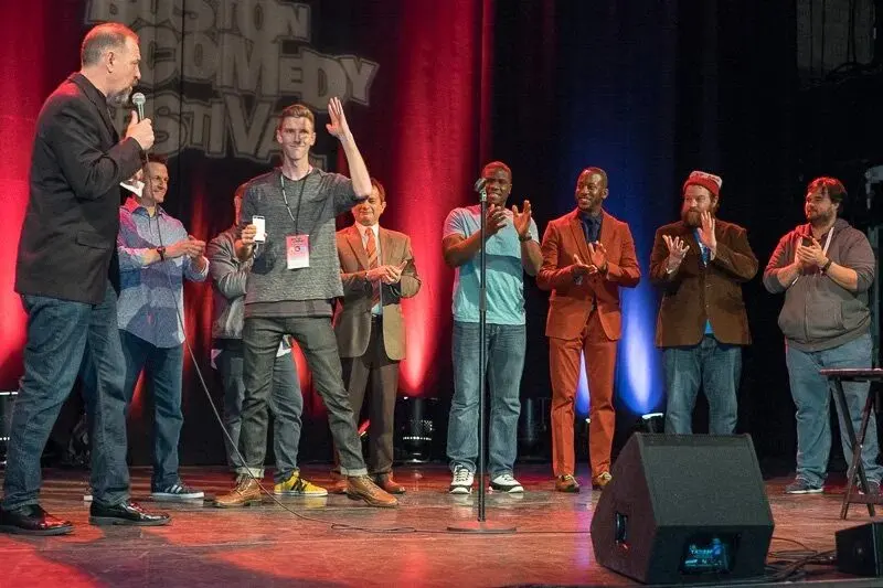 Comedy competition finalists lined up on the festival stage, applauding as the host speaks, with the Boston Comedy Festival