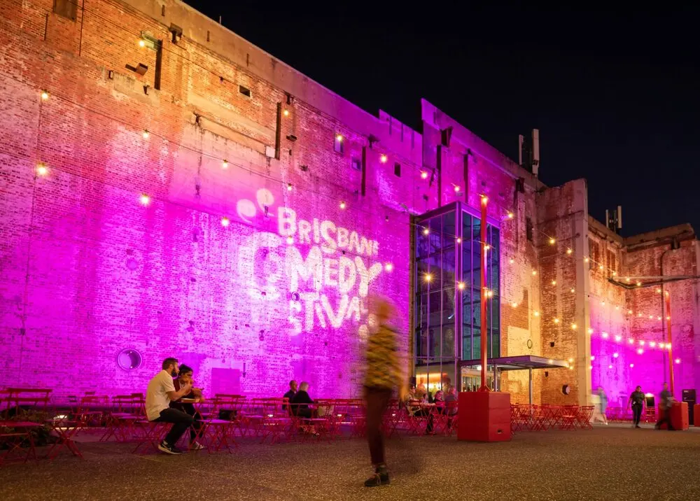 The Brisbane Powerhouse at night lit pink with the festival's name projected on its brick wall, people at tables outside.