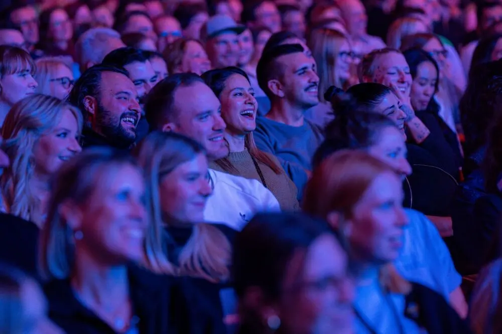 A smiling, laughing audience in purple light at the festival's 1LIVE Comedy-Nacht XXL in the LANXESS arena.