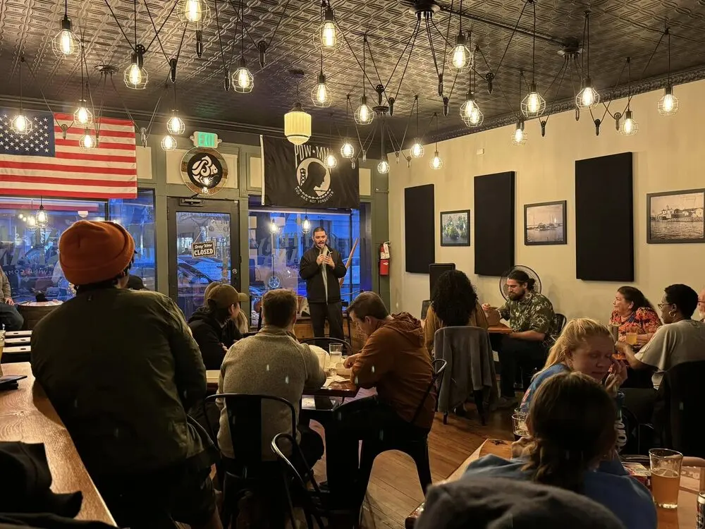A comedian at the mic by the front windows of a tap room full of guests at tables under hanging bulbs during the HarCo