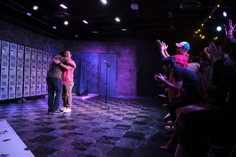 Two comedians hugging on a purple-lit stage while the front row cheers and claps at Laugh After Dark ComedyFest