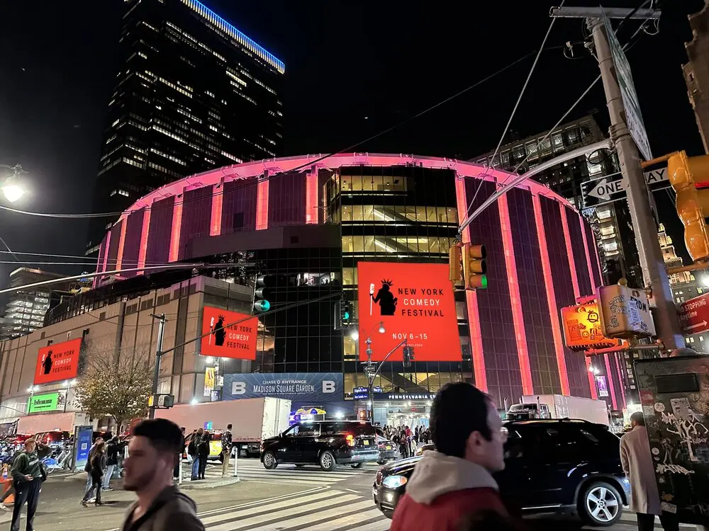 Madison Square Garden lit up at night with New York Comedy Festival banners on its walls and crowds crossing Eighth Avenue