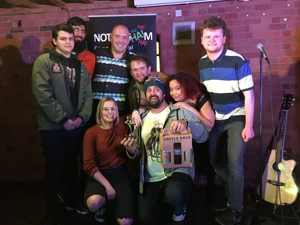 A group of smiling comedians posing with a trophy in front of the festival's banner.