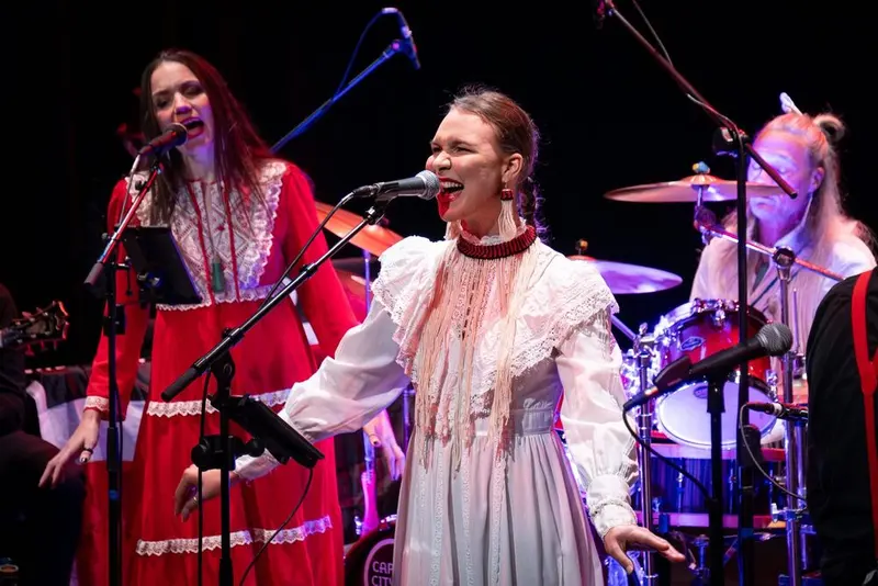Singers in folk dresses performing at microphones with a band on stage at an NZ Fringe 2026 show.
