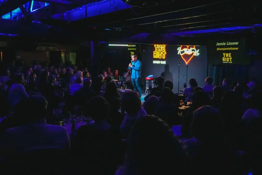 A full room watching a comedian on a small stage under blue light, The Riot sign glowing behind him.
