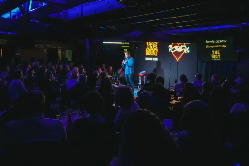 A full room watching a comedian on a small stage under blue light, The Riot sign glowing behind him.