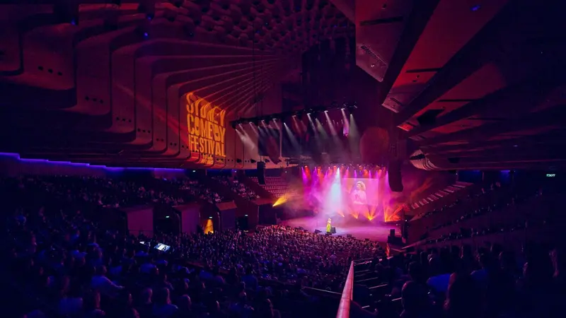 A huge audience in a concert hall facing a pink-lit stage, the Sydney Comedy Festival sign glowing above.