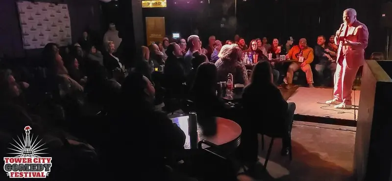 A comedian on a small stage facing a full, dimly lit room of Tower City Comedy Festival guests at their tables