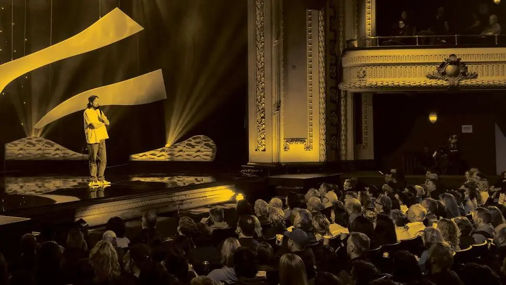 A comedian on a lit stage in a gilded theatre, gold-tinted, with a full audience in the stalls and balcony.