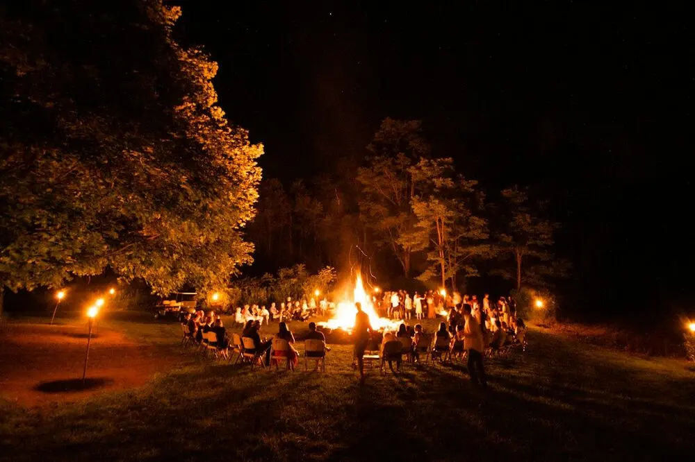 Big campfire at night ringed by folding chairs and tiki torches, crowd as backs and blurred shapes
